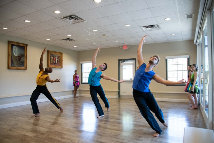 three male dancers in bright colors move on a smooth wooden floor to their right are paintings and to their right and left are women dancer waiting for their cue to come forward